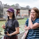 Two women laugh beside a lily pond in front of old gray-walled houses with red lanterns