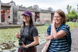 Two women laugh beside a lily pond in front of old gray-walled houses with red lanterns