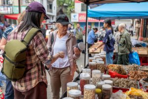 A woman in a cap holds up a jar at a stall of dried goods as another woman gestures toward it